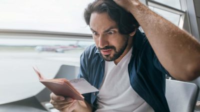 a man sitting in an aeroplane seat holds the top of his head as he looks at his airline ticket with an annoyed, angry expression on his face.
