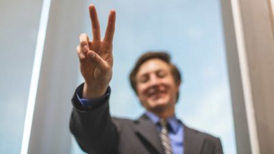 A businessman in soft-focus holds two fingers in the air in the foreground of the shot as he stands smiling in the background against a clear sky.