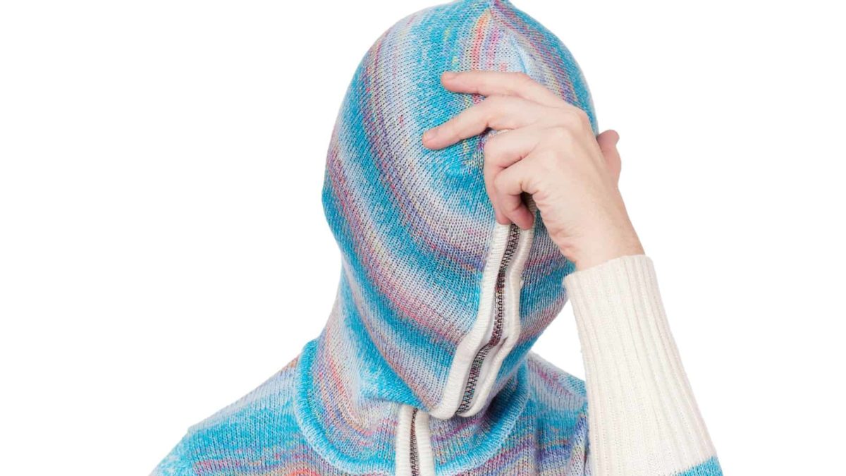 a person zips their jumprer completely over their head, covering their face and holds a hand to their head as if in despair.