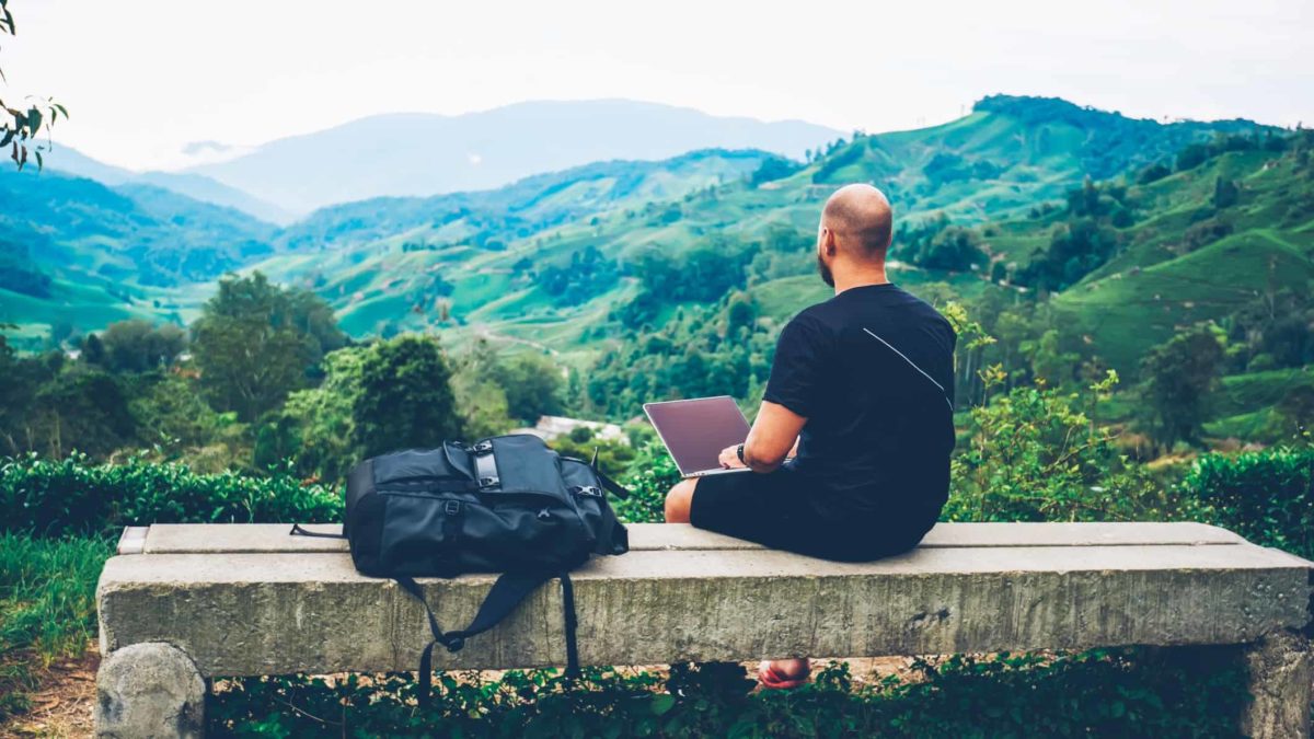 A man sits on a bench atop a mountain with a laptop, making investments with a green ESG mind.