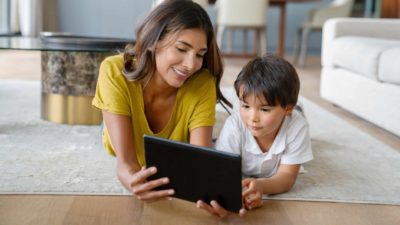 A mother and her young son are lying on the floor of their lounge sharing a tech device.