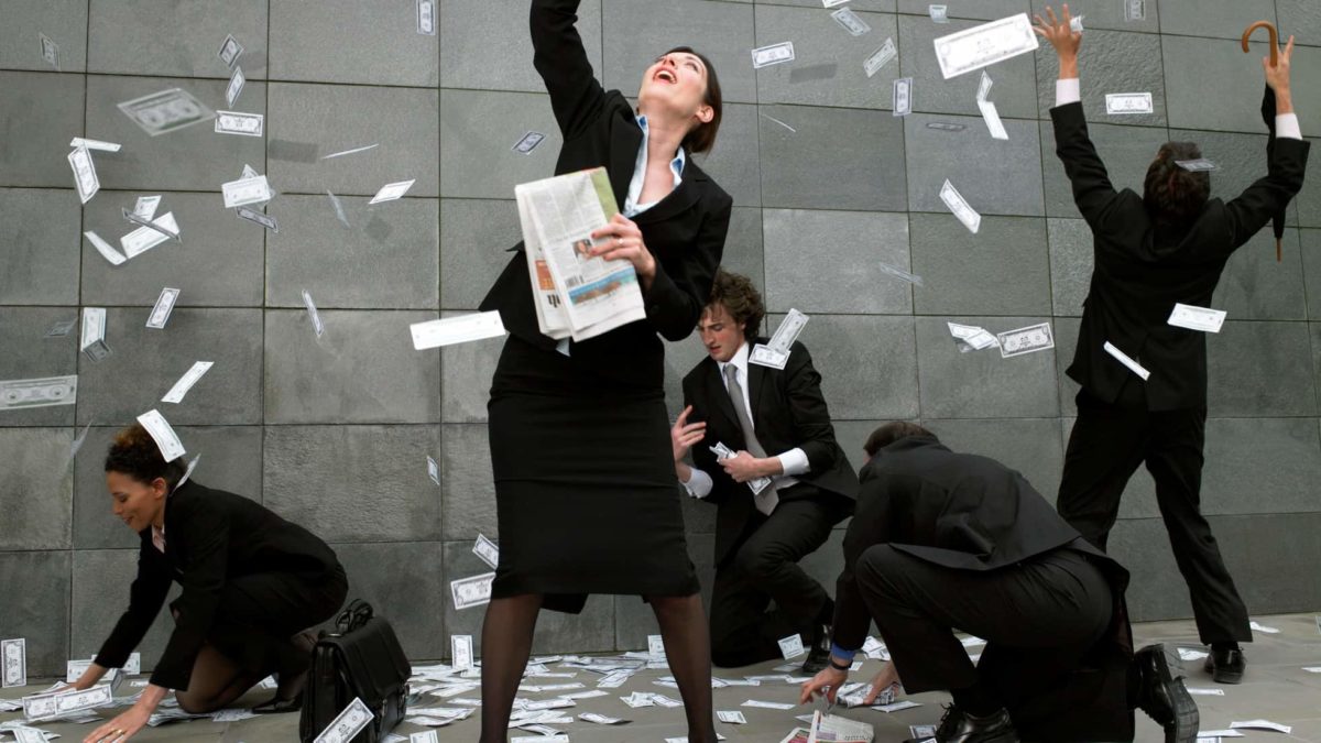 A group of five people dressed in black business suits scrabble in a flurry of banknotes that are whirling around them, some in the air, others on the ground as some of them bend to pick up the money.