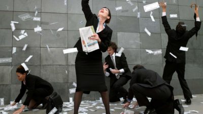 A group of five people dressed in black business suits scrabble in a flurry of banknotes that are whirling around them, some in the air, others on the ground as some of them bend to pick up the money.