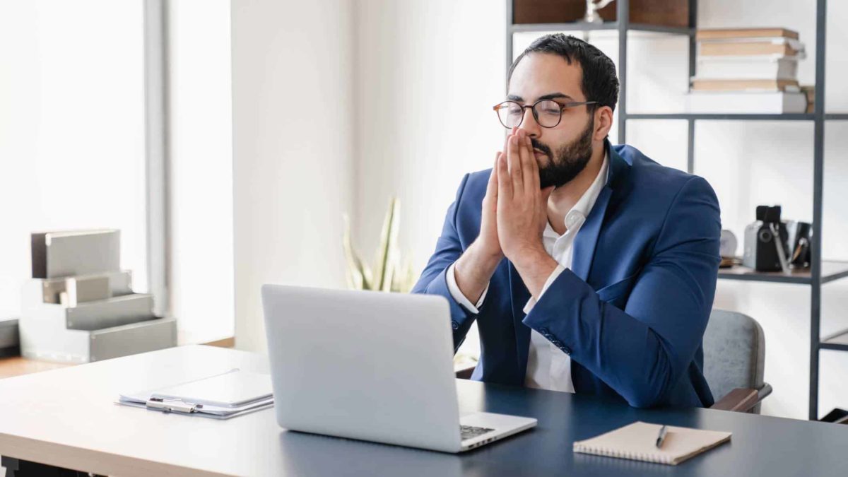 A male investor wearing a white shirt and blue suit jacket sits at his desk looking at his laptop with his hands to his chin, waiting in anticipation.