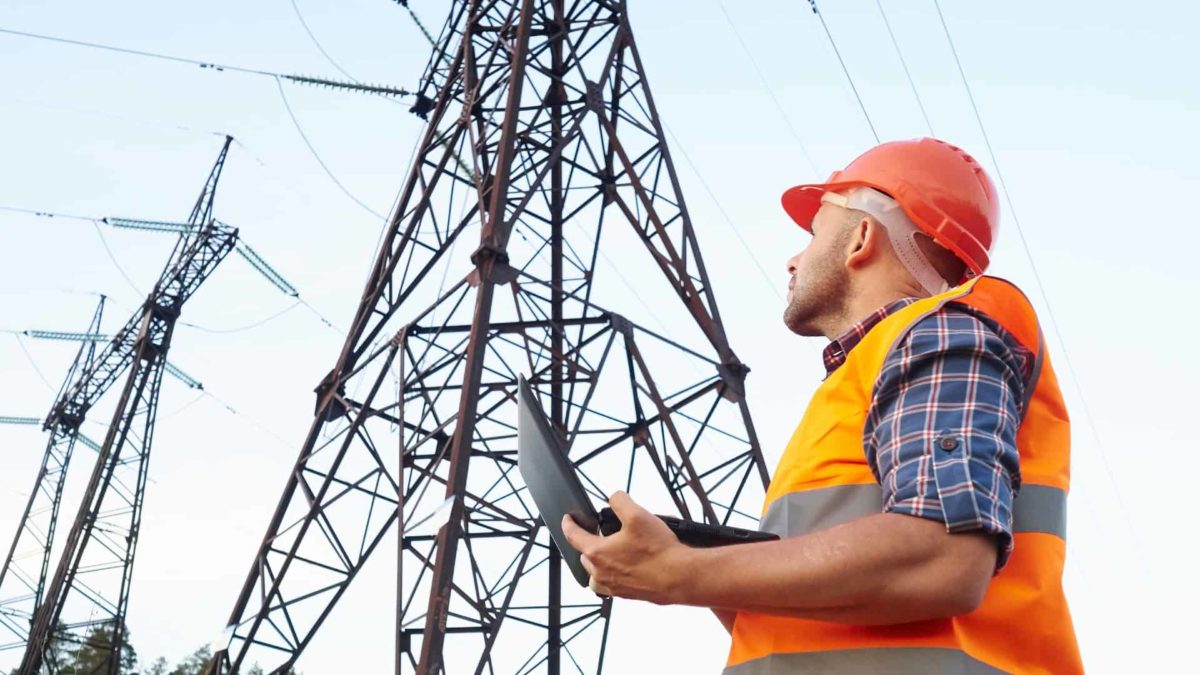 A male electricity worker in hard hat and high visibility vest stands underneath large electricity generation towers as he holds a laptop computer and gazes up at the high voltage wires overhead.