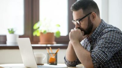 A man sits nervously at his computer with his mouth resting against his hands clasped in front of him as he stares at the screen of his computer on a home desk.