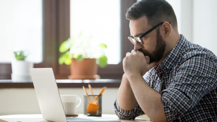 A man sits nervously at his computer with his mouth resting against his hands clasped in front of him as he stares at the screen of his computer on a home desk.