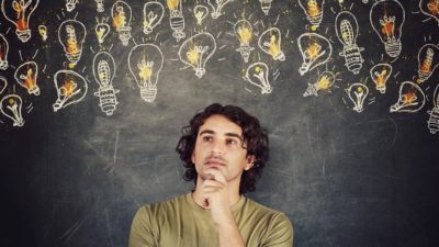 A young man looks like he his thinking holding his hand to his chin and gazing off to the side amid a backdrop of hand drawn lightbulbs that are lit up on a chalkboard.