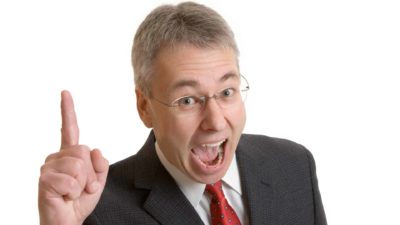 A man in a business suit and a tie leans forward with an excited, wide-mouthed expression and holds up one finger to the camera as if indicating the number one.