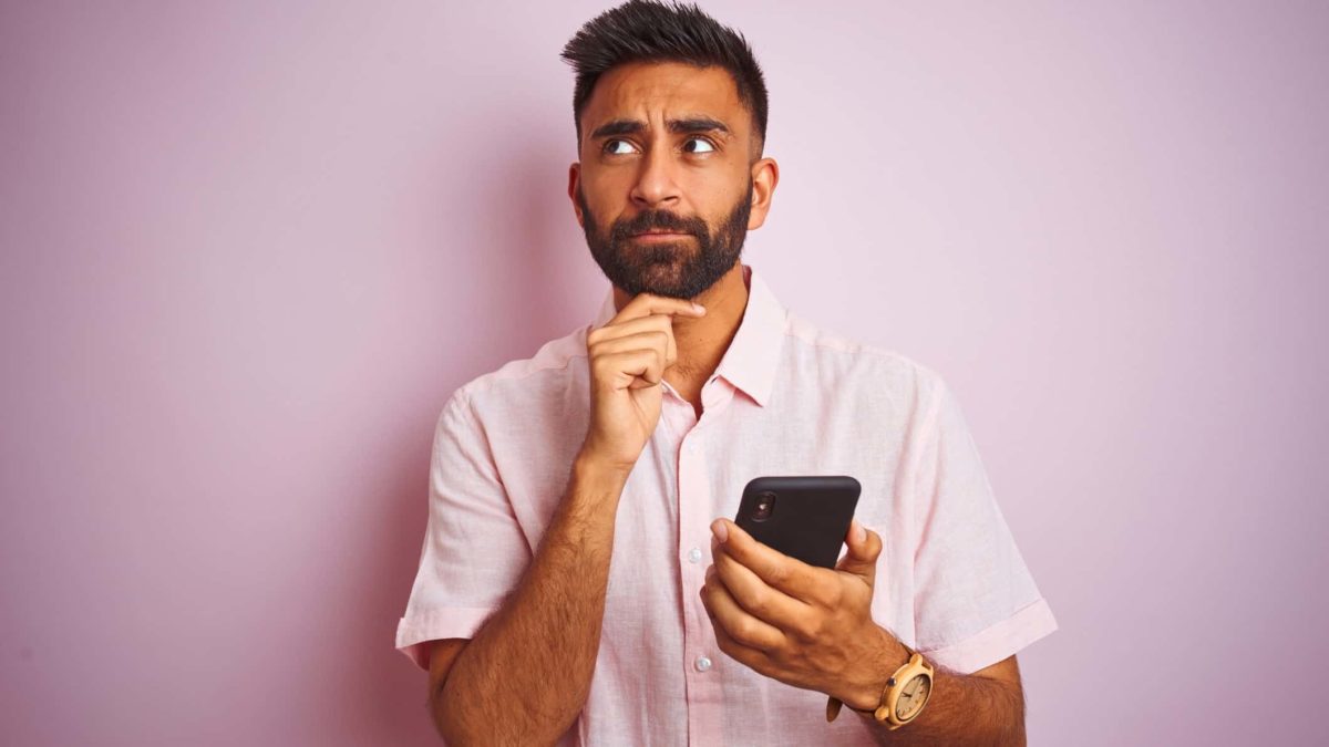 A man casually dressed looks to the side in a pensive, thoughtful manner with one hand under his chin, holding a mobile phone in his hand while thinking about something.