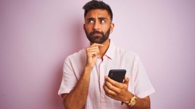A man casually dressed looks to the side in a pensive, thoughtful manner with one hand under his chin, holding a mobile phone in his hand while thinking about something.
