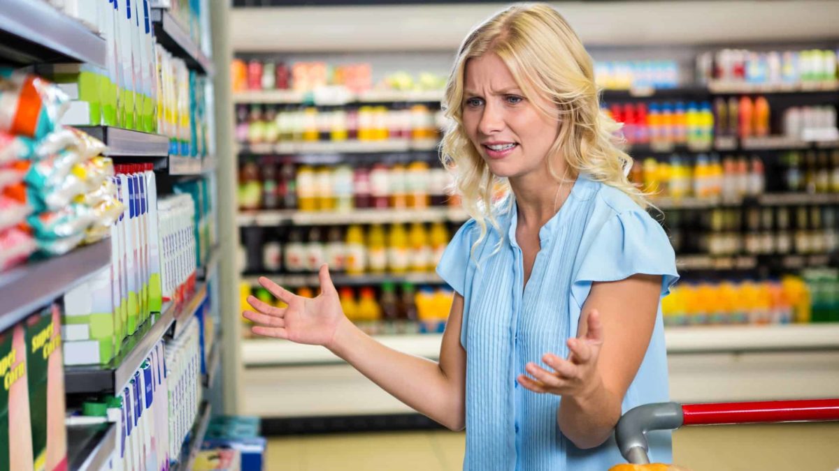 A frustrated young woman shopper holds her hands up with a pained, annoyed expression on her face as she stands next to her trolley in a grocery store and examines the stock offerings on the shelf in front of her.