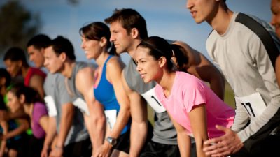 A view of competitors in a running event, some wearing number bibs, line up together on a starting line looking ahead as if to start a race.
