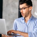 A man in his 30s holds his laptop and operates it with his other hand as he has a look of pleasant surprise on his face as though he is learning something new or finding hidden value in something on the screen.