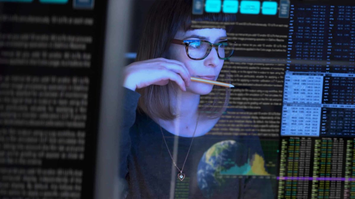 A young woman with glasses holds a pencil to her lips as she is surrounded by the reflection of data as though she is being photographed through a glass screen project with digital data.