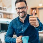 A man holding a cup of coffee puts his thumb up and smiles while at laptop.