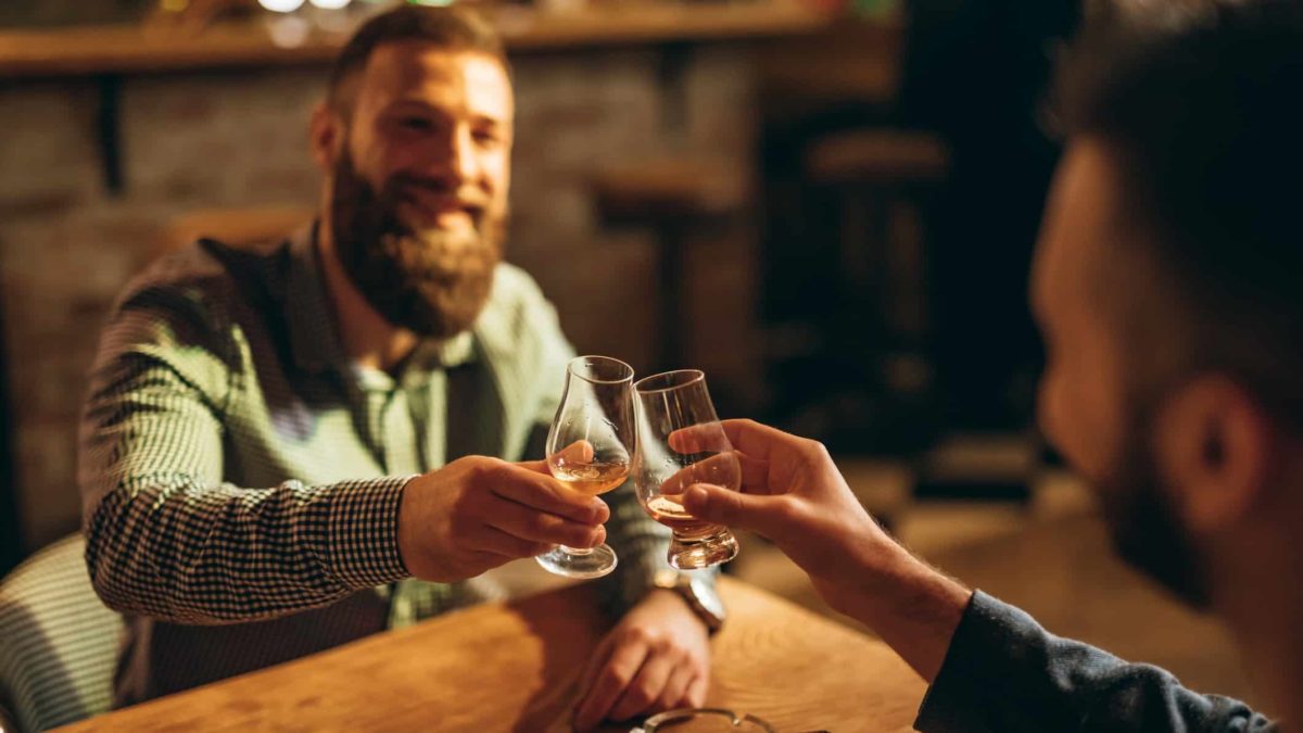 Two men clink whisky glasses while sitting at a table.