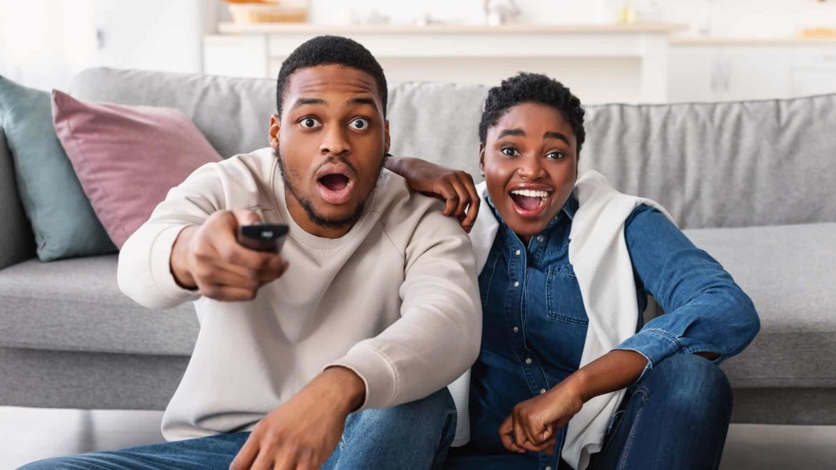 A couple stares at the tv in shock, one holding the remote up ready to press.