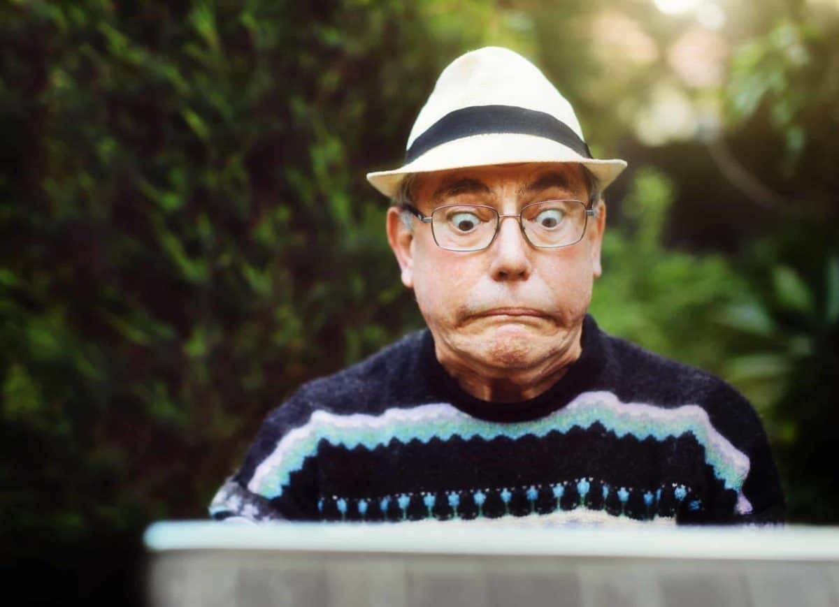 A man wearing a blue jumper and a hat looks at his laptop with a distressed and fearful look on his face.
