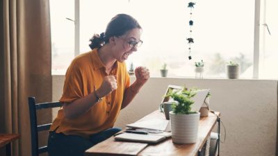 A woman is very excited about something she's just seen on her computer, clenching her fists and smiling broadly.