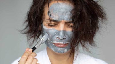 A woman grimaces as she applies a clay beauty mask to her face.