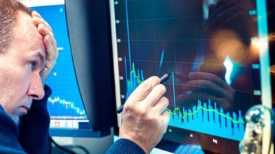 a man sits with his head in his hand, looking quite dejected, as he holds a rubber tipped pen on the screen of a computer showing a graph trending downwards.