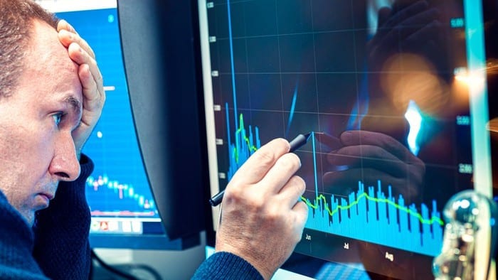 a man sits with his head in his hand, looking quite dejected, as he holds a rubber tipped pen on the screen of a computer showing a graph trending downwards.