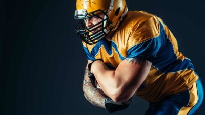 A man in full American NFL playing kit crouches over with his arms across his chest in a defensive stance against a dark background.
