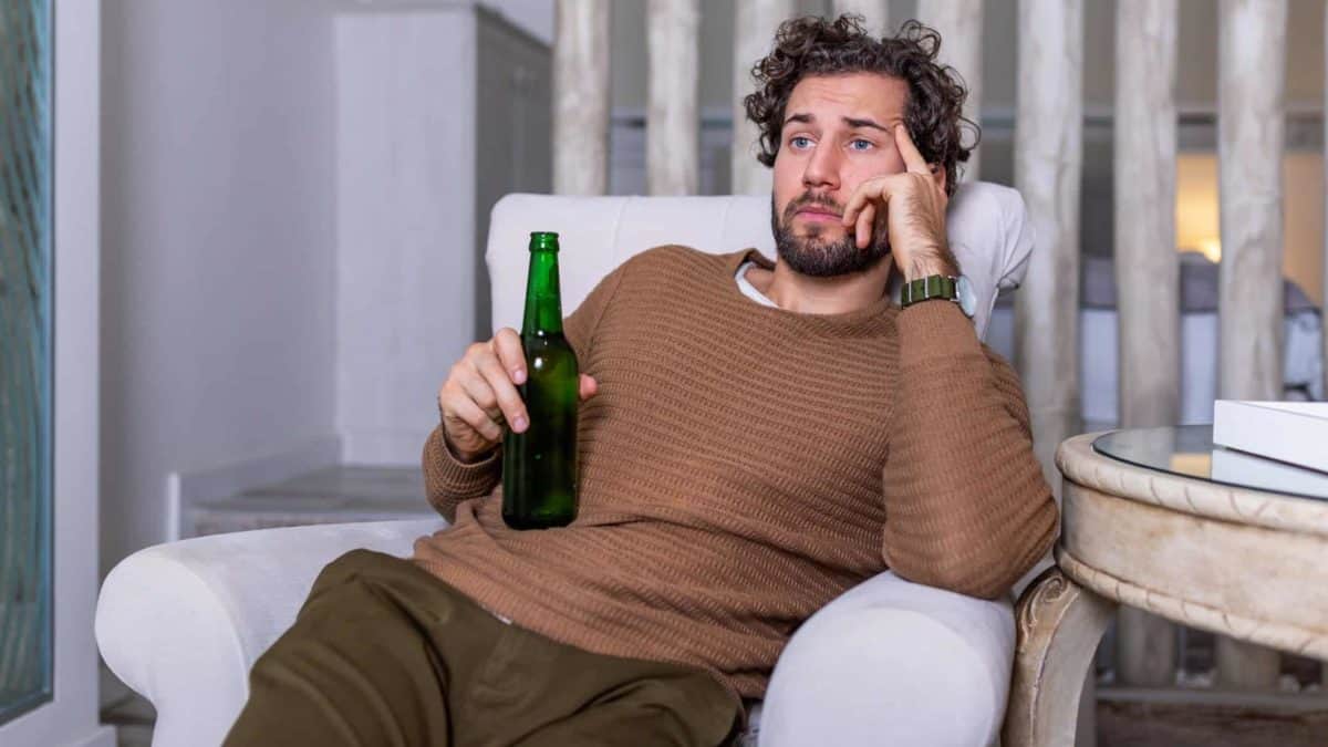A young man holds a small bottle of beer as he slumps sadly on one elbow in a comfortable chair with his head propped in his hand and staring into space with a dejected look on his face.